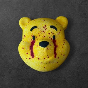 May include: A yellow bear-shaped bath bomb featuring black eyes, a black nose, and arched eyebrows. The surface is decorated with red splatters and red streaks running down from the eyes, creating a distinct contrast against the bright yellow.