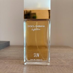 Op de afbeelding: Een rechthoekige glazen parfumfles gevuld met amberkleurige vloeistof. De fles heeft een witte dop, een goudkleurige hals en de witte tekst Dolce & Gabbana light blue SUN EAU DE TOILETTE op de voorzijde van het glas.