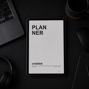 Pode incluir: Uma configuração de espaço de trabalho preto com um laptop, tablet, fones de ouvido, mouse, xícara de café e uma pequena planta. O tablet exibe a palavra "PLANNER" e "undated". Os itens estão dispostos em uma superfície preta.