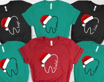 May include: A collection of t-shirts in various colors, including black, teal, and red. Each shirt features a white tooth graphic wearing a Santa hat. Names like "Angel", "Pamela", and "Julia" are written on the shirts, along with the word "Dentist".