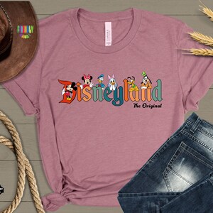 Puede incluir: Una camiseta rosa con un logotipo de Disneyland de estilo vintage que presenta a Mickey Mouse, Minnie Mouse, Donald Duck, Goofy, Pluto y Daisy Duck. El texto debajo del logotipo dice "The Original".