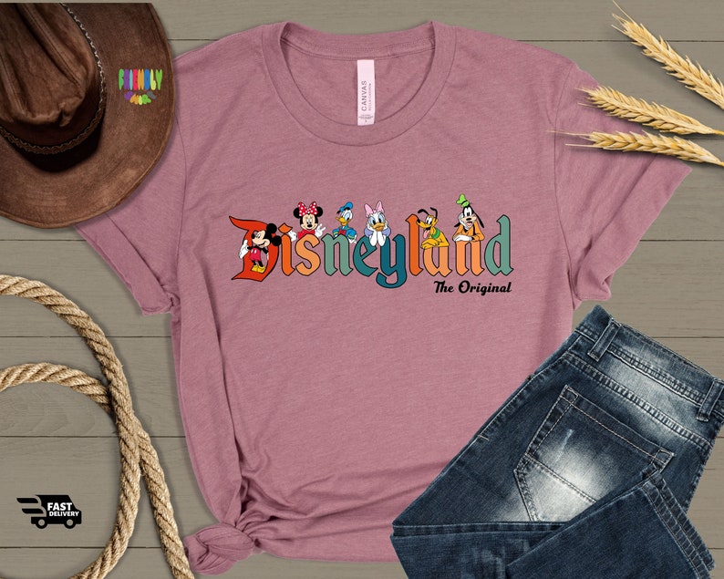 Puede incluir: Una camiseta rosa con un logotipo de Disneyland de estilo vintage que presenta a Mickey Mouse, Minnie Mouse, Donald Duck, Goofy, Pluto y Daisy Duck. El texto debajo del logotipo dice "The Original".