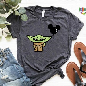 May include: Dark gray t-shirt featuring a cartoon image of a green alien character holding a black Mickey Mouse balloon. The shirt has rolled-up sleeves and is paired with blue jeans and sandals. The text "FRIENDLY SHOPS" is visible in the upper right corner.