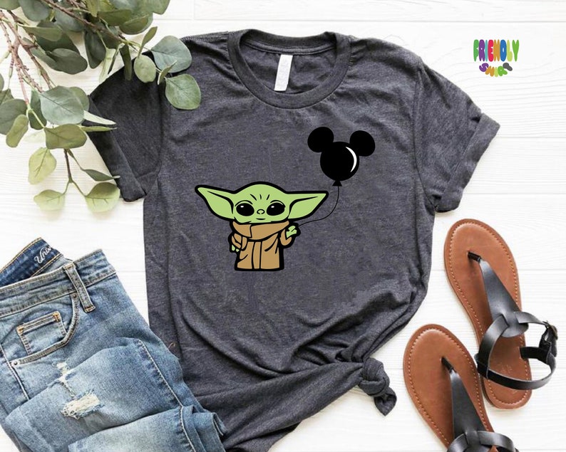May include: Dark gray t-shirt featuring a cartoon image of a green alien character holding a black Mickey Mouse balloon. The shirt has rolled-up sleeves and is paired with blue jeans and sandals. The text "FRIENDLY SHOPS" is visible in the upper right corner.