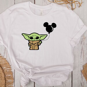 May include: White t-shirt featuring a cartoon illustration of a green alien-like character holding a black Mickey Mouse balloon. The character is wearing a brown coat and scarf. The design is centered on the shirt.