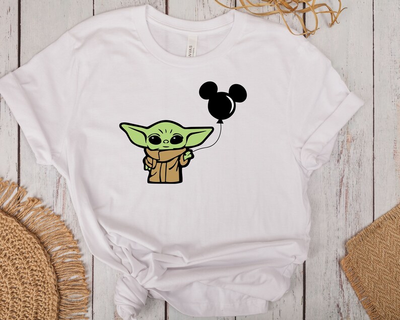 May include: White t-shirt featuring a cartoon illustration of a green alien-like character holding a black Mickey Mouse balloon. The character is wearing a brown coat and scarf. The design is centered on the shirt.