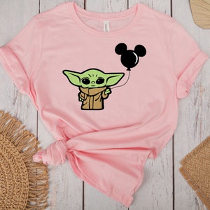 May include: A light pink t-shirt featuring a cartoon character holding a black Mickey Mouse balloon. The character is green with large eyes and wears a brown cloak. The t-shirt is made of soft material.