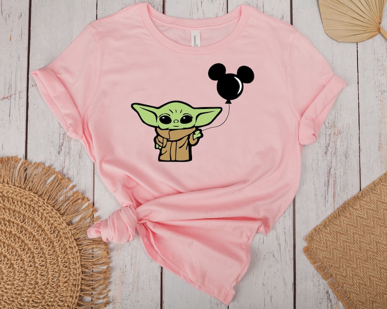 May include: A light pink t-shirt featuring a cartoon character holding a black Mickey Mouse balloon. The character is green with large eyes and wears a brown cloak. The t-shirt is made of soft material.
