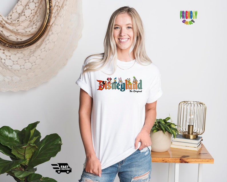 Puede incluir: Camiseta blanca con un gr&aacute;fico colorido del logotipo de Disneyland y personajes. El texto dice "Disneyland The Original".