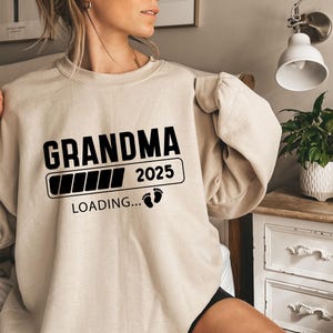 May include: A beige crewneck sweatshirt featuring black text that reads GRANDMA, followed by a loading bar graphic, the year 2025, and the text LOADING... accompanied by two small black baby footprint icons centered on the chest area.