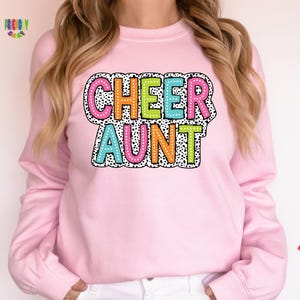 May include: A light pink long-sleeve sweatshirt featuring the text CHEER AUNT in colorful, polka-dotted block letters. The letters are outlined in black and set against a white background with a black speckled pattern, creating a bold graphic design.