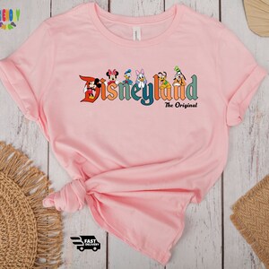 Puede incluir: Camiseta rosa con un gr&aacute;fico del logotipo de Disneyland y varios personajes de Disney. El texto "The Original" est&aacute; debajo del logotipo.