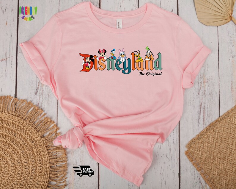 Puede incluir: Camiseta rosa con un gr&aacute;fico del logotipo de Disneyland y varios personajes de Disney. El texto "The Original" est&aacute; debajo del logotipo.