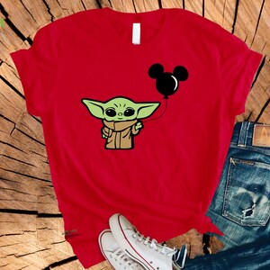May include: Red t-shirt featuring a cartoon image of Grogu, also known as Baby Yoda, holding a black Mickey Mouse balloon. The design is in a cartoon style. The shirt is paired with blue jeans and white sneakers.