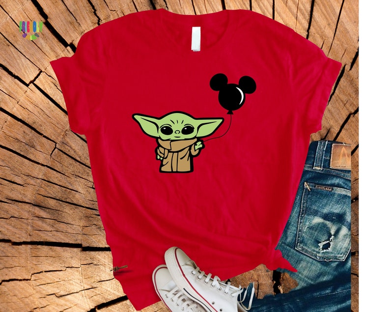 May include: Red t-shirt featuring a cartoon image of Grogu, also known as Baby Yoda, holding a black Mickey Mouse balloon. The design is in a cartoon style. The shirt is paired with blue jeans and white sneakers.