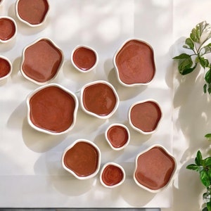 Puede incluir: Una colección de 11 pequeños cuencos de cerámica de color terracota de diversas formas y tamaños. Los cuencos están dispuestos sobre una superficie blanca.