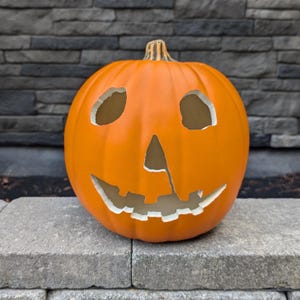 May include: A carved orange pumpkin with a smiling face, featuring triangular eyes and a jagged mouth. The pumpkin is a classic Halloween decoration, perfect for seasonal displays. The pumpkin is sitting on a gray stone surface.