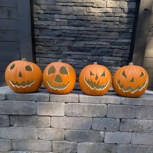 May include: Four orange artificial pumpkins arranged in a row, each featuring a unique carved jack-o'-lantern face with cutouts for eyes, noses, and jagged mouths. The pumpkins have smooth, rounded surfaces and are positioned on a gray stone ledge.