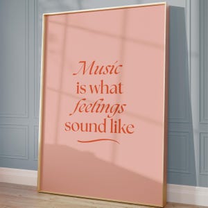 May include: A rectangular wall art print in a thin gold frame featuring a dusty pink background with the dark orange text "Music is what feelings sound like" written in a serif font, centred with a decorative horizontal line underneath.