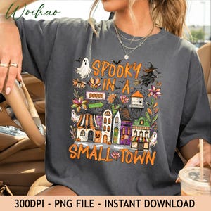 May include: A charcoal grey t-shirt featuring a colourful Halloween-themed graphic with the text "SPOOKY IN A SMALL TOWN" and "90001". The design includes illustrations of a ghost, a witch on a broomstick, a water tower, and various buildings.