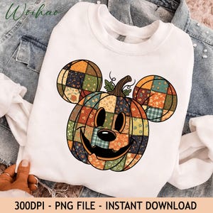 May include: A white crewneck sweatshirt featuring a large graphic of a pumpkin shaped like a mouse head. The pumpkin has a patchwork design with various floral and plaid patterns in orange, green, and navy blue, with a carved face and stem.