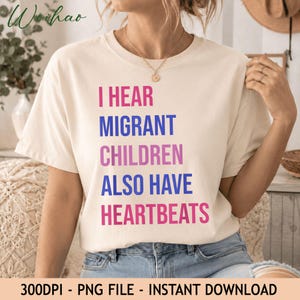 Puede incluir: Una camiseta de manga corta de color crema con el texto I HEAR MIGRANT CHILDREN ALSO HAVE HEARTBEATS impreso en líneas alternas de letras mayúsculas rosas y azules. La camiseta tiene cuello redondo y un corte relajado.