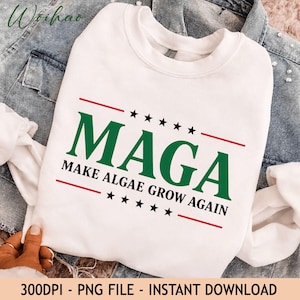 May include: A white crewneck sweatshirt featuring the green text MAGA and the black text MAKE ALGAE GROW AGAIN. The design includes two horizontal red lines and two rows of five black stars positioned above and below the text elements.