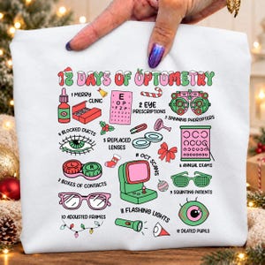 May include: A white sweatshirt featuring a colourful graphic titled 12 Days of Optometry. The design includes twelve illustrations representing eye care themes, such as eye charts, contact lens cases, glasses, and medical equipment, all in a festive palette.