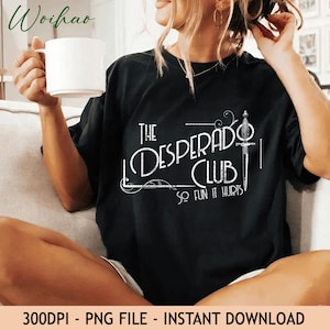 May include: A black short-sleeved t-shirt featuring white graphic text that reads The Desperado Club So Fun It Hurts, accompanied by a detailed illustration of a dagger. The design includes decorative flourishes and lines surrounding the text.
