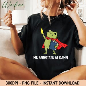 May include: A black t-shirt featuring a green frog wearing a red cape, holding a yellow highlighter and a blue book with colorful page markers. The text "WE ANNOTATE AT DAWN" is printed in white below the frog graphic on the front of the shirt.