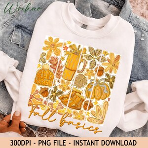 May include: A white crewneck sweatshirt featuring a graphic print of autumn-themed items including a knit hat, a mug, boots, pumpkins, acorns, and flowers in shades of yellow and orange, with the text Fall basics written in cursive script.