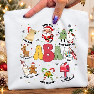 May include: A white sweatshirt featuring a colorful graphic with the text ABA in large letters. Surrounding the text are Christmas-themed illustrations including Santa, a snowman, a reindeer, a gingerbread man, and candy canes with labels.