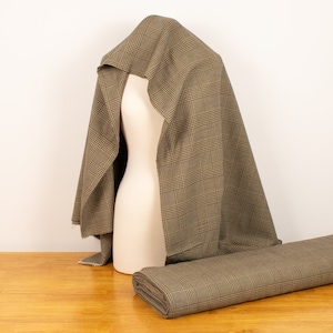 May include: A brown and beige plaid fabric draped over a dress form. The fabric is rolled up and placed on a wooden table.