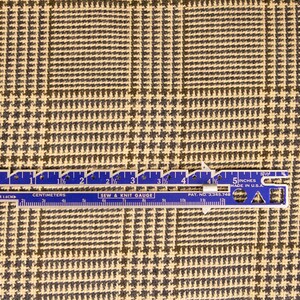 Puede incluir: Un primer plano de una tela de pata de gallo beige y negra con una regla que mide 5 pulgadas. La regla es azul con marcas blancas y el texto "5 inches made in U.S.A."