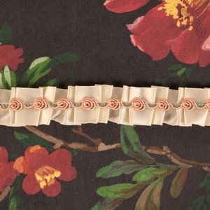 May include: A close-up of a cream-colored satin ribbon trim with pink rosettes and green accents. The trim is on a dark brown background with a floral pattern.
