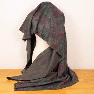May include: A gray shawl with a patterned design in shades of teal and burgundy is draped over a mannequin. The shawl has a fringed edge and is displayed on a wooden surface.