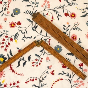 May include: Two wooden folding rulers with brass hinges. The rulers are laid out in a V shape on a white fabric with a floral pattern. The rulers have markings in inches.