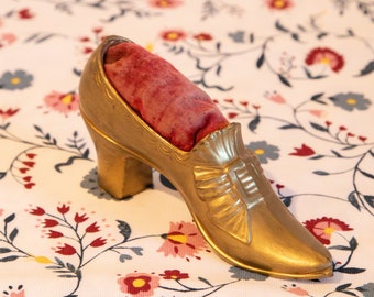 Puede incluir: Un alfiletero en forma de zapato de color dorado con un interior de terciopelo rojo. El alfiletero está sobre una tela blanca con un patrón floral.