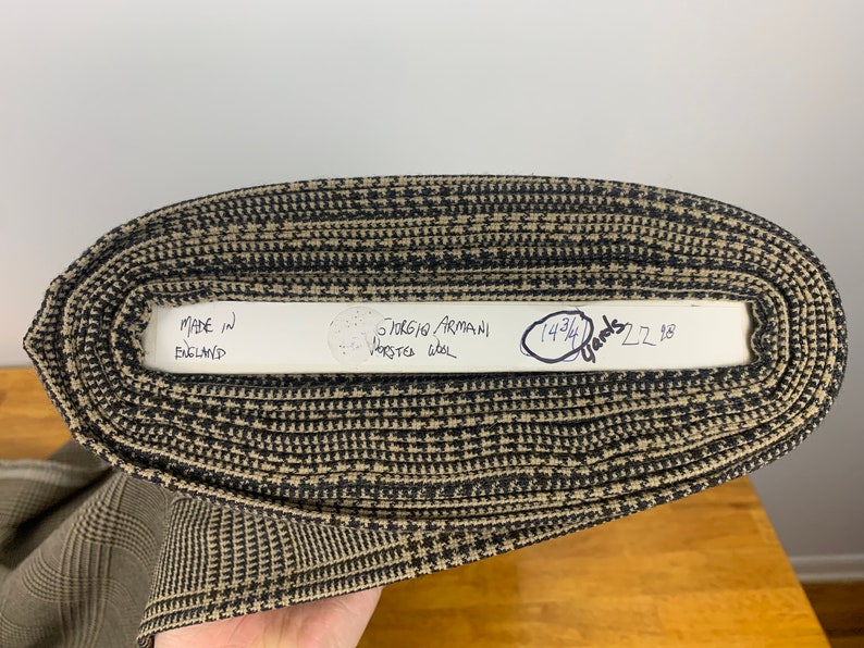 Puede incluir: Primer plano de un rollo de tela con estampado marr&oacute;n y negro con una etiqueta blanca que dice "Made in England" y "Giorgio Armani Worsted Wool" con una nota manuscrita que dice "14 3/4 Yards 22.98".