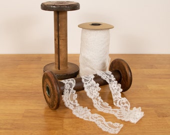 Puede incluir: Dos carretes de madera con cinta de encaje blanco enrollada alrededor. Los carretes son marrones y tienen un acabado marrón oscuro. El encaje es delicado y tiene un patrón floral.