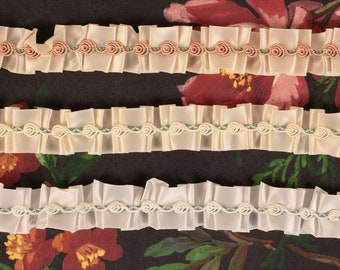 Puede incluir: Tres filas de cinta de raso de color marfil con detalles verdes y rosas. La cinta tiene un borde fruncido y un patrón repetitivo de pequeñas rosetas.