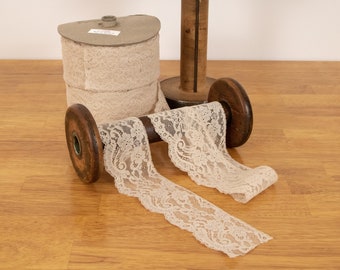 Puede incluir: Dos carretes de cinta de encaje de color blanco roto enrollados alrededor de carretes de madera. El encaje tiene un delicado patrón floral.