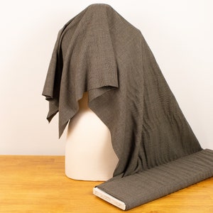 May include: A roll of olive green fabric with a subtle striped pattern, draped over a white mannequin head. The fabric is also laid out flat on a wooden surface, showcasing its texture and design.