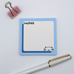 May include: Blue notepad with a white border and the word "Notes" printed in black. A small cartoon cat is in the bottom right corner. A gold pen is lying on a white surface.