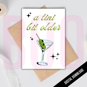 Puede incluir: Una tarjeta de felicitación rosa y blanca con una copa de martini y aceitunas. El texto de la tarjeta dice "a  tini bit older".