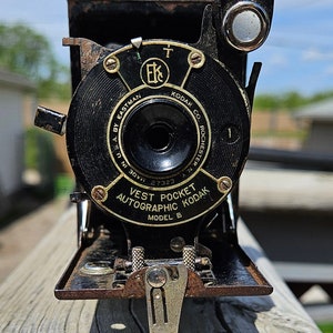 May include: A black vintage Vest Pocket Autographic Kodak camera with a leather case. The camera has a round lens with a black metal rim and a silver metal lever on the bottom. The camera is made in the USA by Eastman Kodak Co. Rochester, NY. The camera has a serial number of 27323.