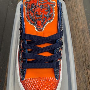 May include: Orange and navy blue embellished sneaker featuring a Chicago Bears logo. The toe and upper are adorned with sparkling rhinestones. The shoe has navy blue laces and a white sole with a red trim.