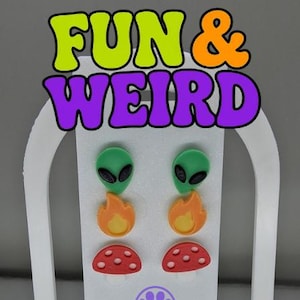 May include: A set of six stud earrings arranged on a white display card with the text FUN & WEIRD at the top and a Purr Prints logo at the bottom. The earrings feature green alien heads, orange flame shapes, and red mushroom caps with white spots.