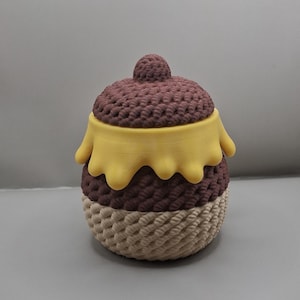 Puede incluir: Un recipiente pequeño y redondo con tapa, que presenta un patrón texturizado similar a una cuerda en marrón oscuro en la parte superior y beige claro en la inferior. Un diseño liso y amarillo, como gotas, rodea la sección superior.