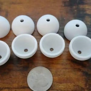May include: Eight white, hemispherical, hollow objects with a small circular hole at the center of each piece. The items are arranged in two rows of four, positioned above a single silver-colored United States quarter coin for scale reference.